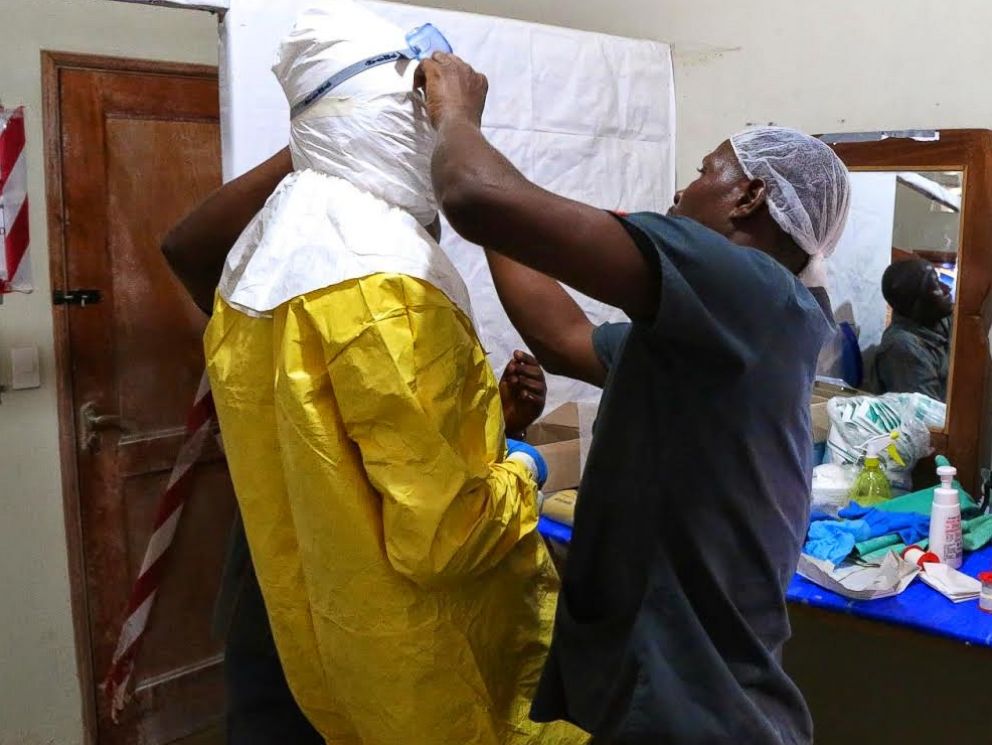 PHOTO: Three people help Dr. Besser put on protective gear outside the ELWA Ebola Isolation Unit in Monrovia, Liberia.