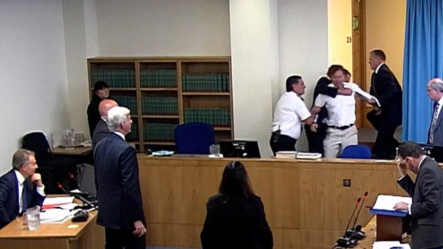 PHOTO: Britains former Prime Minister Tony Blair, left, looks on, May 28 2012, as a protester, top center in white shirt, is restrained by officials after he burst in through a secure corridor shouting, "This man should be arrested for war crimes!".