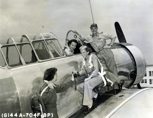 Photo of three women and a man with a plane. One woman is sitting in the pilot's seat, talking to the two women crouching by the side of the plane. The man is standing next to the women, smiling broadly at the camera.