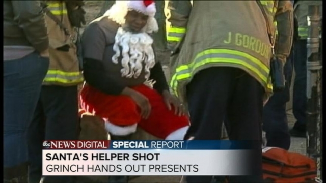 Man in Santa Claus suit shot in back at toy giveaway, Grinch volunteer aids in giveaway.